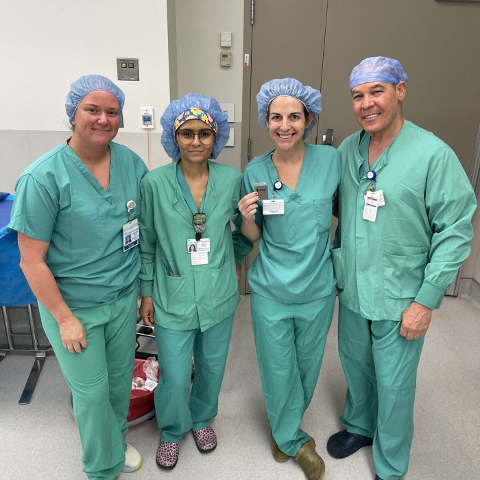 Four medical professionals in green scrubs and blue surgical caps stand together in a hospital room, smiling at the camera.