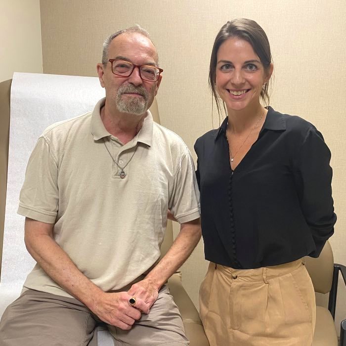An older man sits on a medical exam table next to a young woman standing and smiling in an examination room.