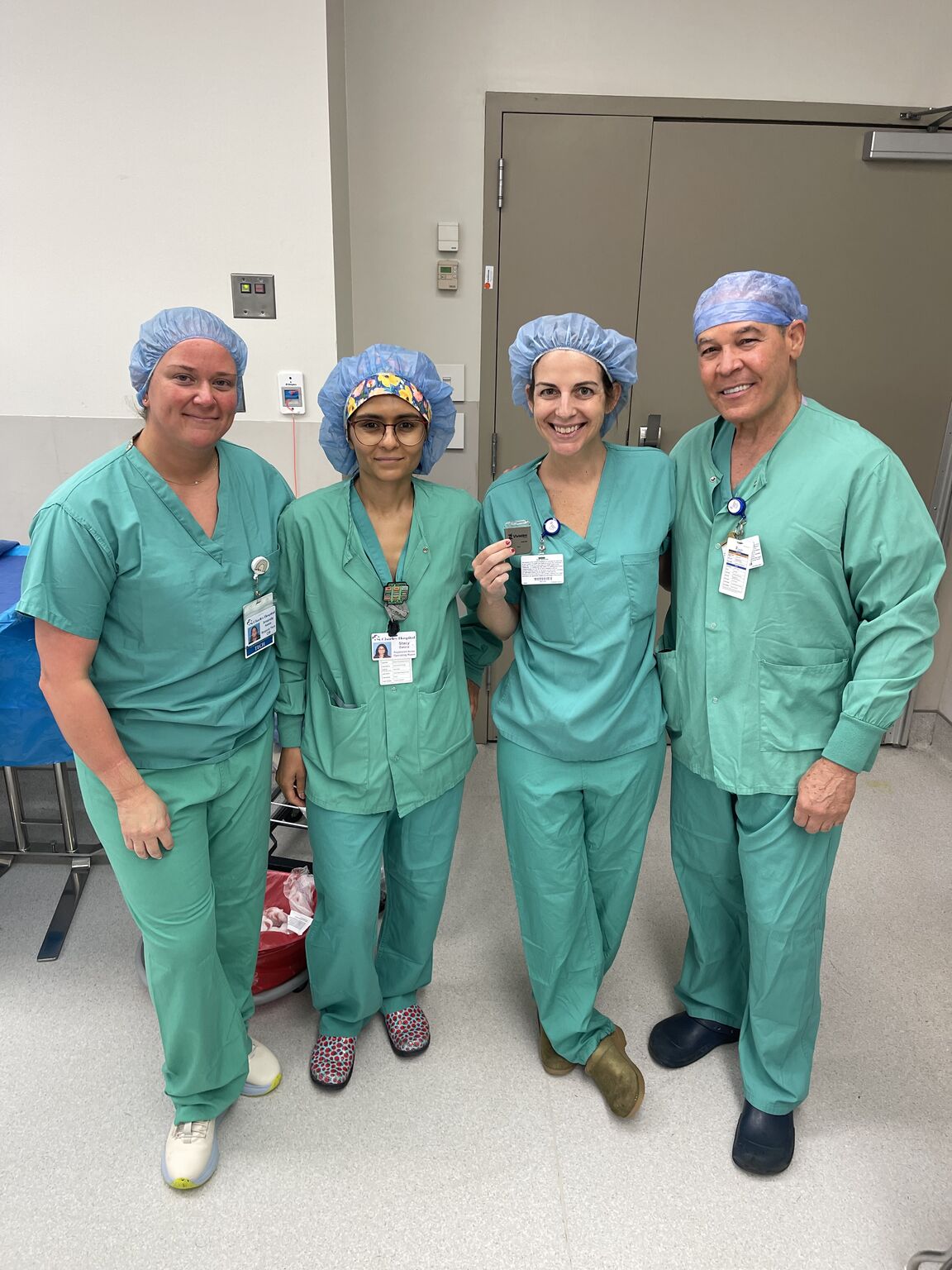 Four medical professionals in green scrubs and blue surgical caps stand together in a hospital room, smiling at the camera. Four medical professionals in green scrubs and blue surgical caps stand together in a hospital room, smiling at the camera