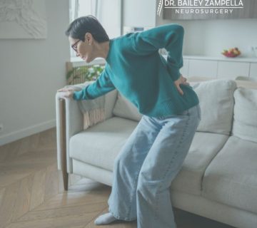 A person in a teal sweater and jeans is standing up from a couch, holding their lower back in pain  The room is softly lit and there is a logo for Dr  Bailey Zampella Neurosurgery 