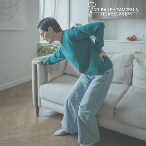 A person in a teal sweater and jeans is standing up from a couch, holding their lower back in pain The room is softly lit and there is a logo for Dr Bailey Zampella Neurosurgery